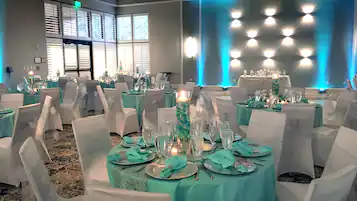 A banquet hall with round tables set for a formal event, featuring teal tablecloths and white chairs with decorative sashes.