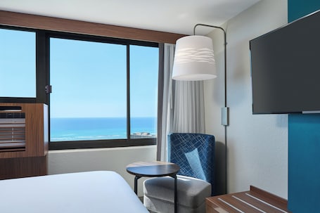 A hotel room with a large window offering a view of the ocean, a bed, a chair, a small table, and a flat-screen TV mounted on the wall.