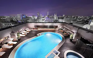 A rooftop pool with lounge chairs and potted plants, overlooking a cityscape at night.