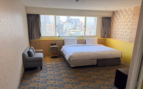 A hotel room with a large bed, a sofa, a small table, and a view of the city through a large window.