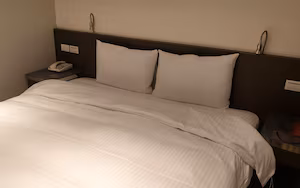 A neatly made bed with white bedding, a bedside telephone, and a nightstand with a lamp.