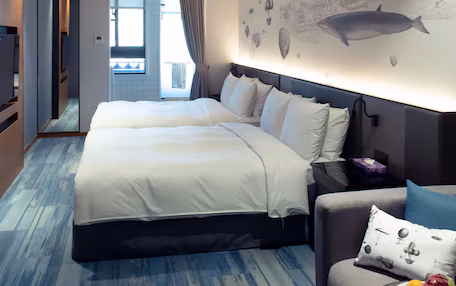 A hotel room with a large bed, a sofa, a dining table with fruit, and a mural of fish and buildings on the wall.