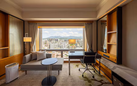 A hotel room with a sofa, a small round table, a desk, and a chair. There is a large window with a city view and mountains in the distance.