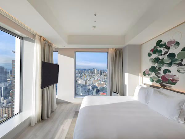 A modern hotel room with a large bed, a flat-screen TV, and a city view through large windows.