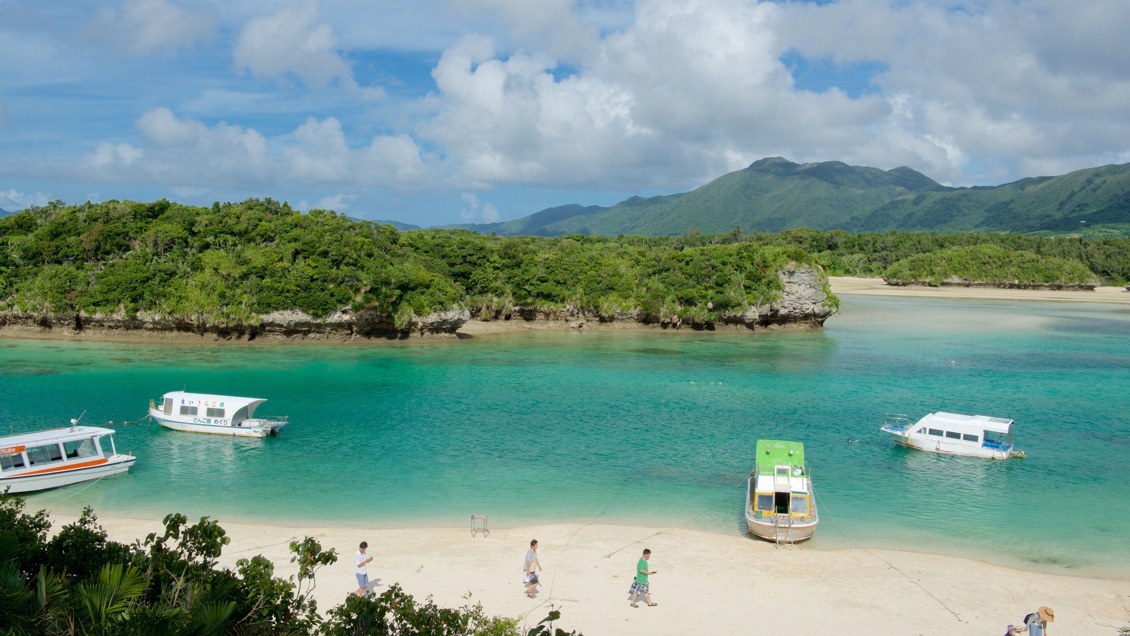 A beach with boats, clear turquoise water, and lush green hills in the background.